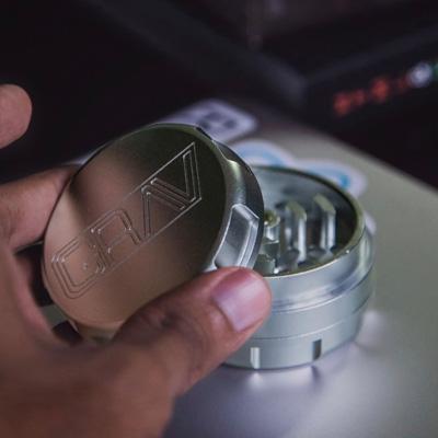 Silver weed grinder sitting on a table with a hand lifting the lid
