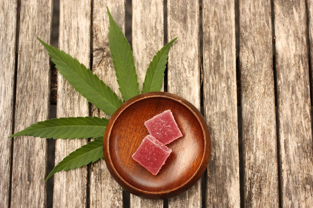  Two gummy candies sitting in a wooden bowl, which is on top of a marijuana leaf, and lying on a wooden porch. 