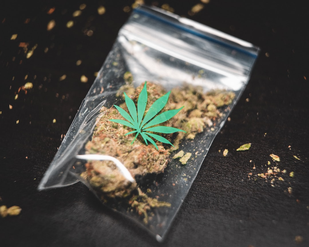  A clear bag of cannabis, with a cannabis leaf printed on it, is sitting on a black surface and surrounded by cannabis crumbs. 