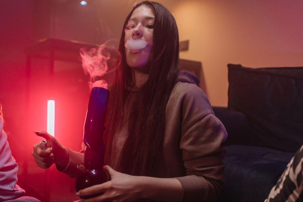  A woman exhales smoke while holding a dark blue glass water pipe under dim pink and purple ambient lighting. 
