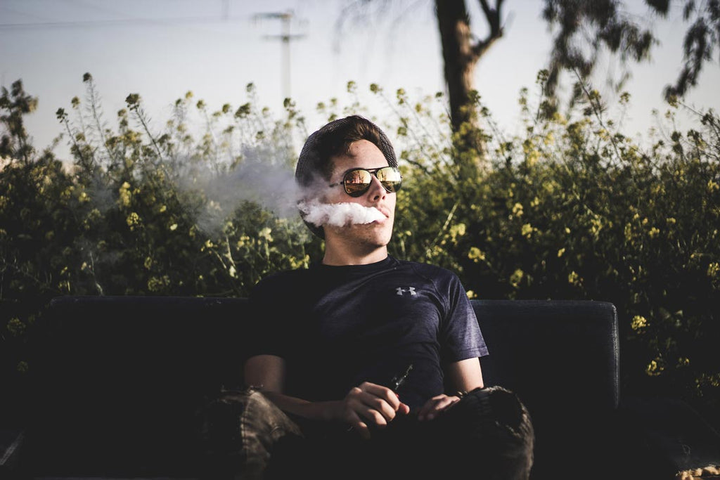  man sitting on a park bench smoking a vape wearing sunglasses 