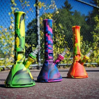 Three colorful and tie-dyed silicone bongs sitting on the ground with trees and a blue sky in the background